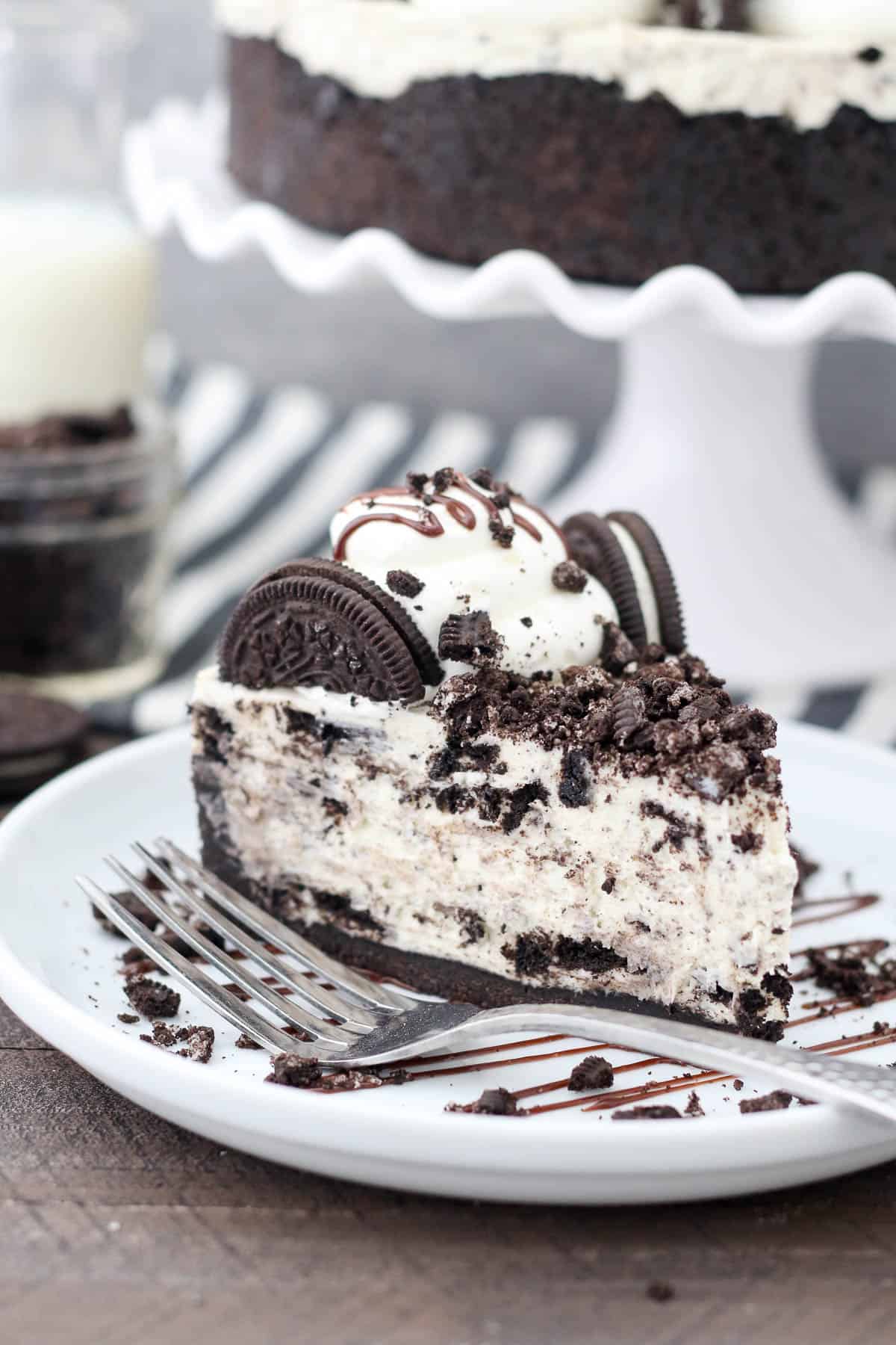 A slice of no-bake Oreo cheesecake on a white plate, topped with Oreos and drizzled with fudge sauce, next to a fork.