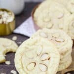 A large almond cookie with 5 sliced almond on top leaning up against a stack of cookie. One of them has a bite taken out of it.