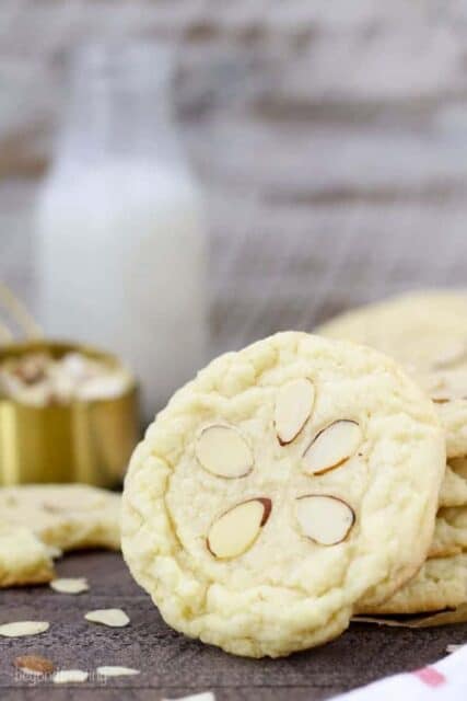 A large almond cookie with 5 sliced almond on top leaning up against a stack of cookies