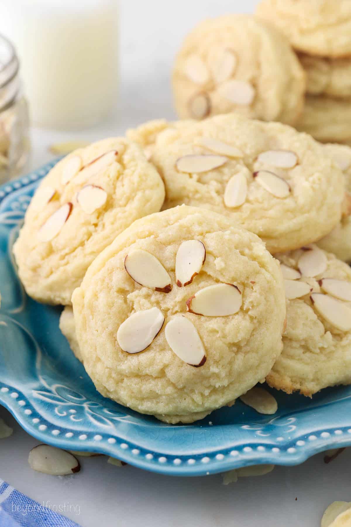 A pile of almond cookies on a blue plate with a glass of milk in the background.