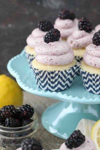 A teal cake stand covered with lemon cupcakes that are topped with a blackberry frosting.