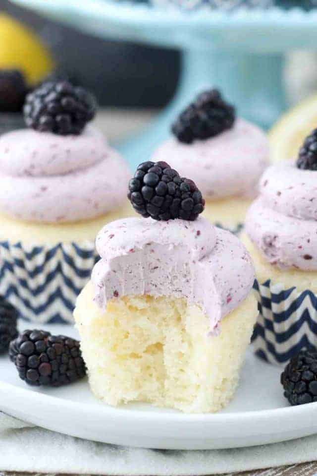A close up shot of a lemon cupcake with a giant bite take out of it. The cupcake is topped with a blackberry buttercream
