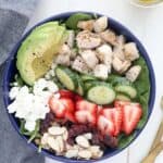 An overhead shot showing the ingredients of the salad including sliced almonds, cranberries, strawberries, goat cheese, cucumbers, chicken, avocado and lettuce. There is a jar of lemon salad dressing in the background and two gold forks.