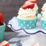 These pretty cupcakes are sitting on a white cake stand. The cupcakes have teal polka dot liners and are topped with a cream cheese frosting and a strawberrty.