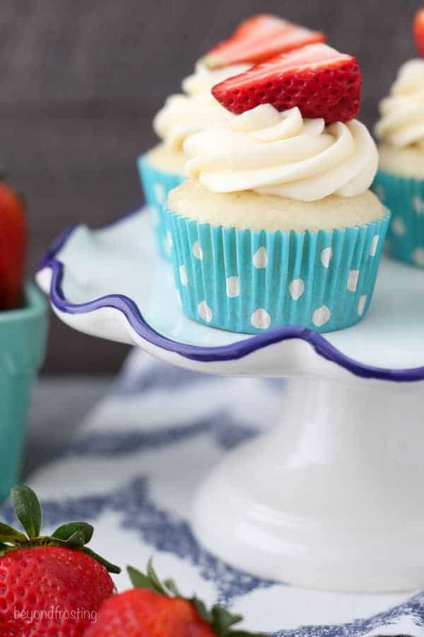 These pretty cupcakes are sitting on a white cake stand with scalloped blue edge. The cupcakes are topped with a pretty white frosting and a strawberry