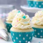A trip of vanilla cupcakes topped with vanilla buttercream. The cupcakes have teal polka dot liners and pink, white and teal sprinkles.