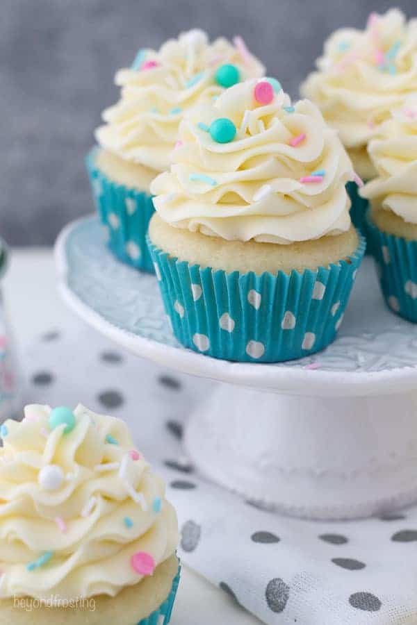 Um pequeno suporte de bolo branco com bolinhos de baunilha que têm forro de polca de teal. Os cupcakes são cobertos com um lindo creme de manteiga de baunilha