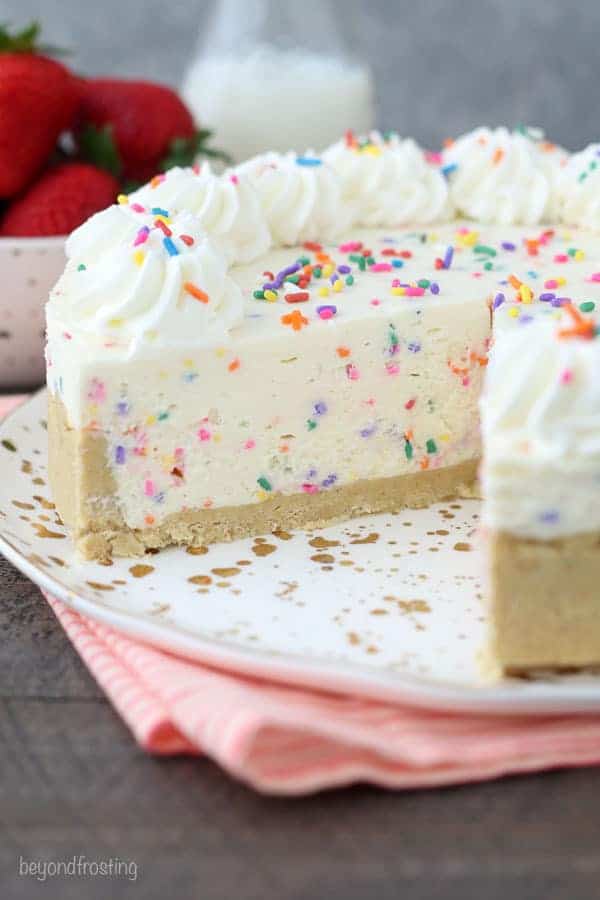 A large no-bake funfetti cheesecake with whipped cream and sprinkles, the cheesecake is sliced and sitting on a gold polka dot plate