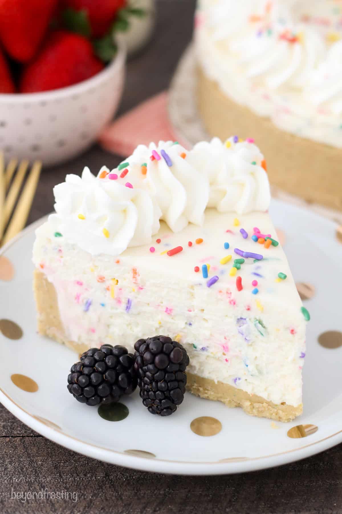 A slice of funfetti cheesecake on a plate, next to two blackberries.