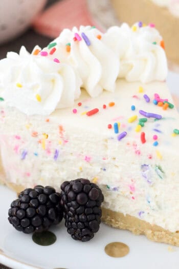 A slice of funfetti cheesecake on a plate, next to two blackberries.