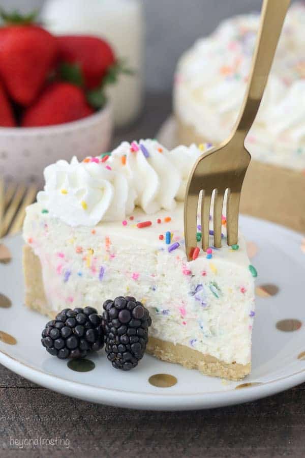 A gold fork sinking into a slice of funfetti cheesecake that is garnished with sprinkles