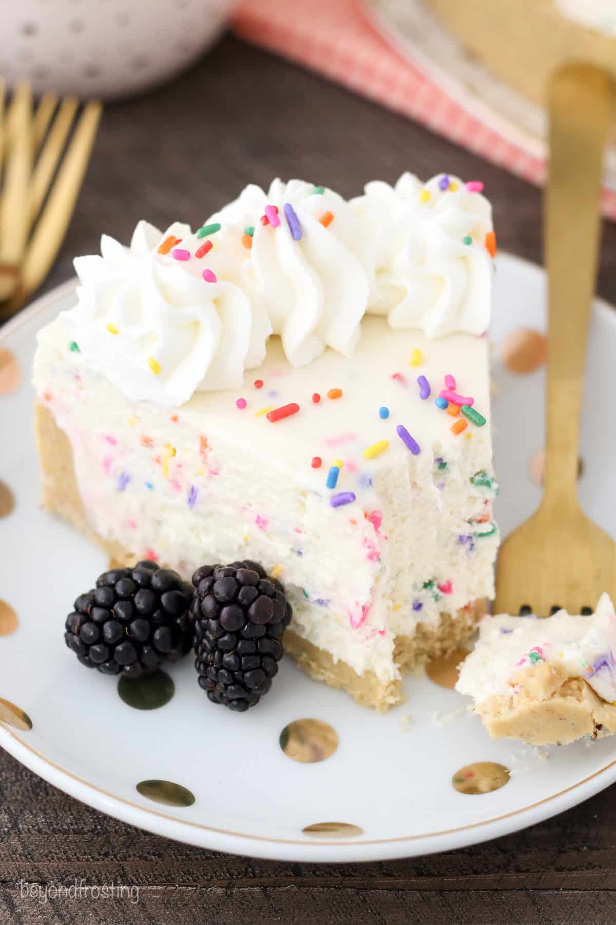 A slice of funfetti cheesecake on a plate with a forkful missing form the end, next to two blackberries.