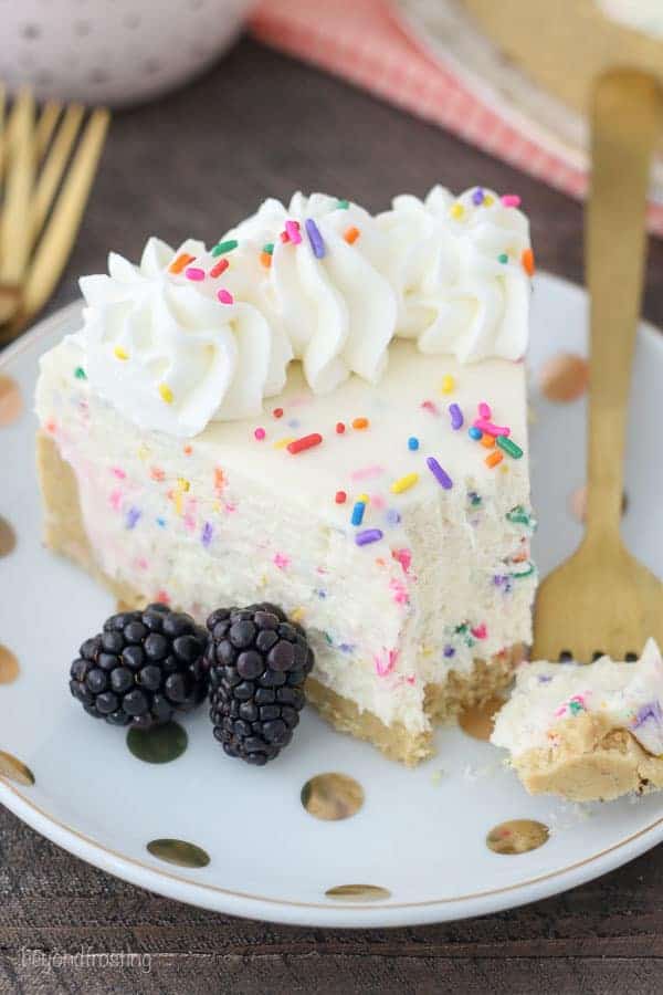 An overhead view of a slice of cheesecake with a bite taken out of it sitting on a gold fork. The cheesecake is garnished with sprinkles and whipped cream and two blackberries.