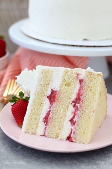 A slide of layered vanilla cake with a strawberry filling and vanilla frosting on a pink plate with a strawberry on top