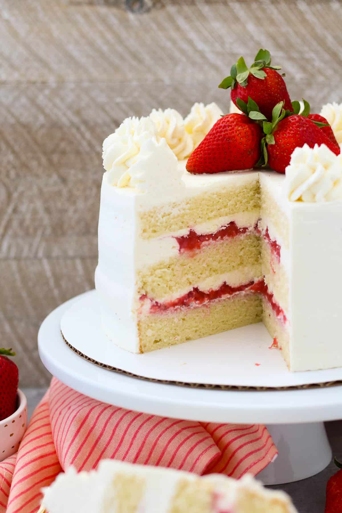 A frosted strawberry mascarpone cake topped with frosting swirls and fresh strawberries on a cake stand, with a slice missing.