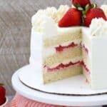 A layered strawberry cake on a white cake plate. The is a big slice missing, showing the inside of the cake and layers of strawberry filling, vanilla cake and frosting.