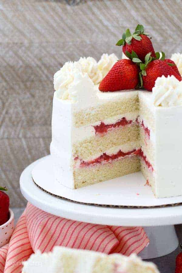A layered strawberry cake on a white cake plate. The is a big slice missing, showing the inside of the cake and layers of strawberry filling, vanilla cake and frosting.