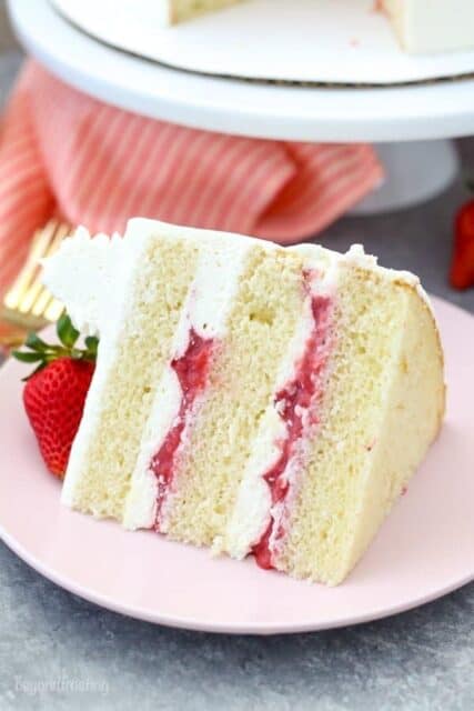 A slide of layered vanilla cake with a strawberry filling and vanilla frosting on a pink plate with a strawberry on top