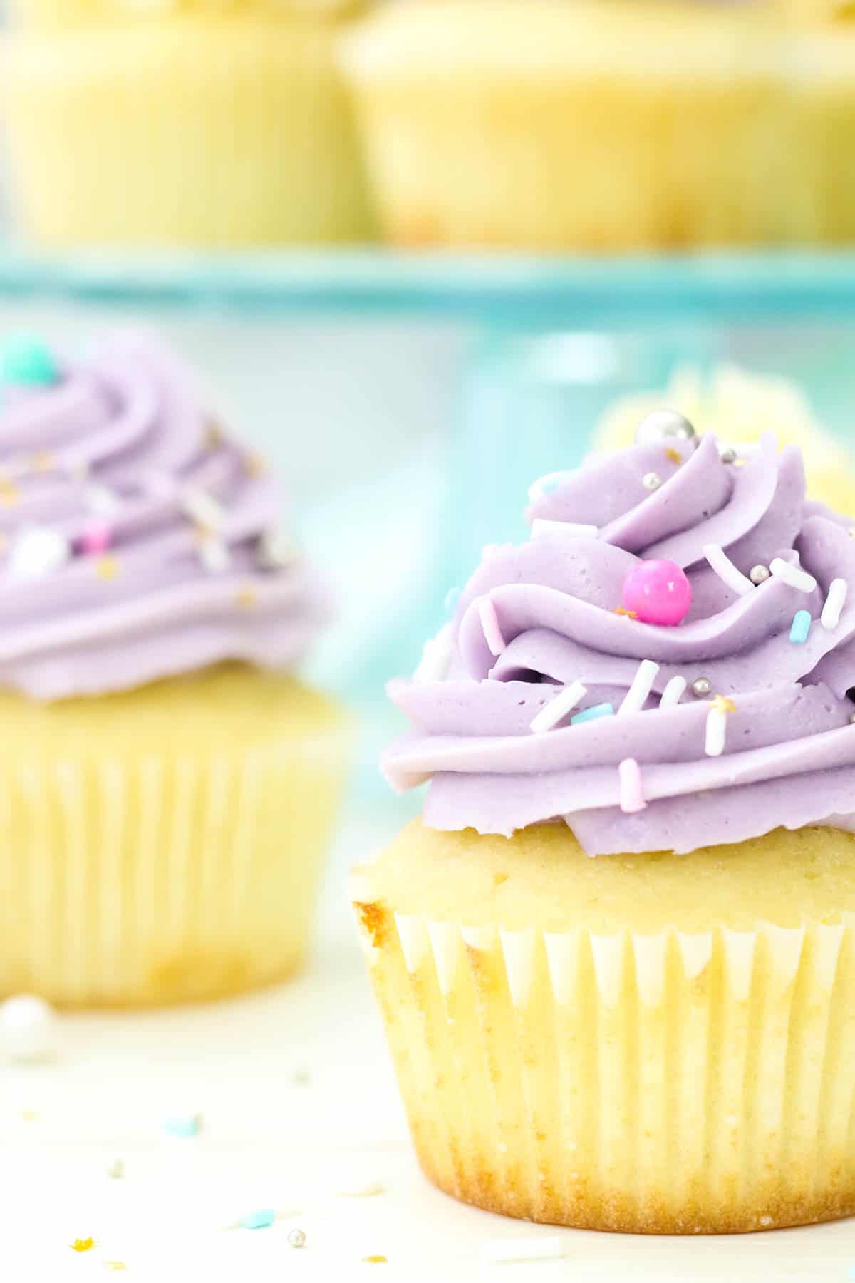 Two vanilla cupcakes frosted with purple Swiss meringue buttercream frosting, with more cupcakes on a cake stand in the background.