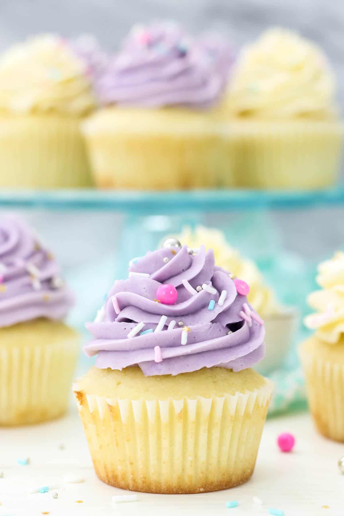 Vanilla cupcakes frosted with purple Swiss meringue buttercream frosting, with more cupcakes on a cake stand in the background.