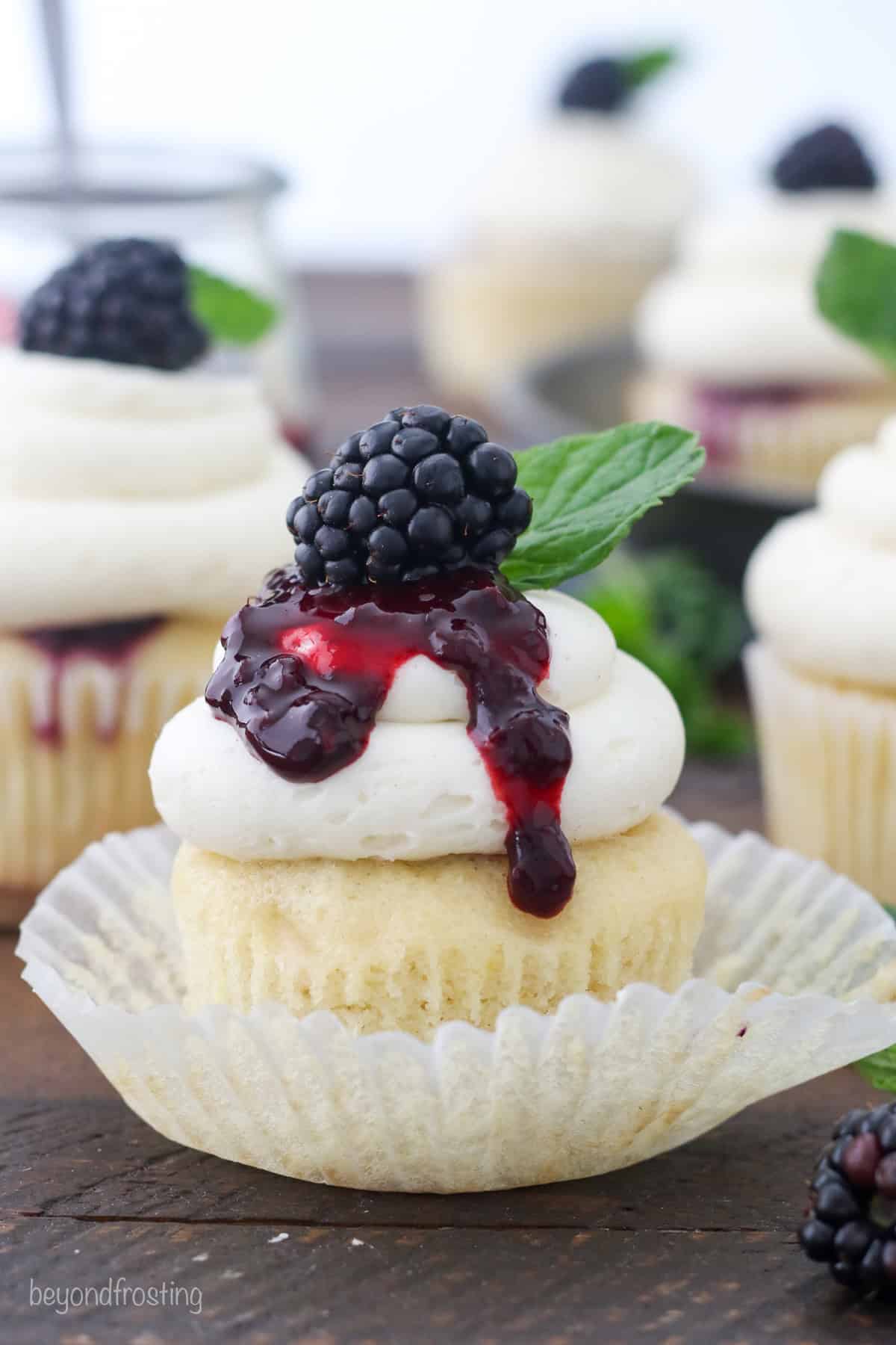 A bourbon smash cupcake inside a cupcake wrapper, topped with vanilla buttercream drizzled with blackberry sauce and garnished with a fresh blackberry and a mint leaf.