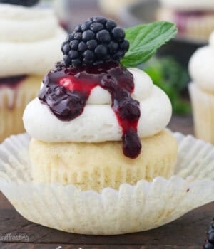 A bourbon smash cupcake inside a cupcake wrapper, topped with vanilla buttercream drizzled with blackberry sauce and garnished with a fresh blackberry and a mint leaf.