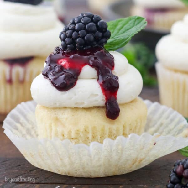 A bourbon smash cupcake inside a cupcake wrapper, topped with vanilla buttercream drizzled with blackberry sauce and garnished with a fresh blackberry and a mint leaf.
