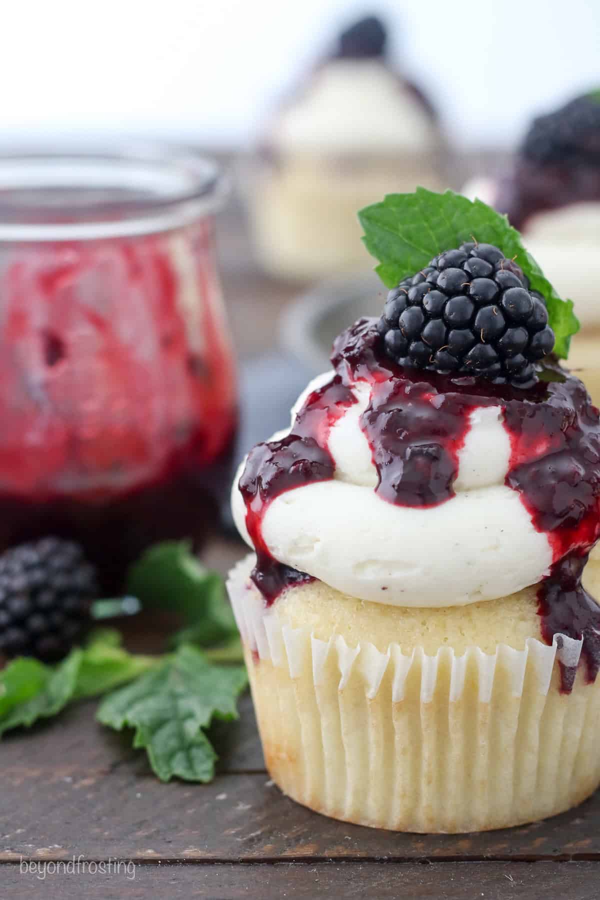 Close up of a bourbon smash cupcake topped with vanilla buttercream drizzled with blackberry sauce, garnished with a fresh blackberry and a mint leaf.
