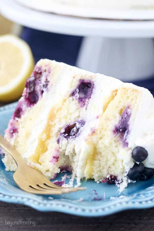 A slice of lemon blueberry cake on a teal cake plate with a few bites taken out of it.