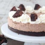 A close up of a brownie batter cheesecake on a white cake plate. The cheesecake is garnished with whipped cream and chopped brownies.