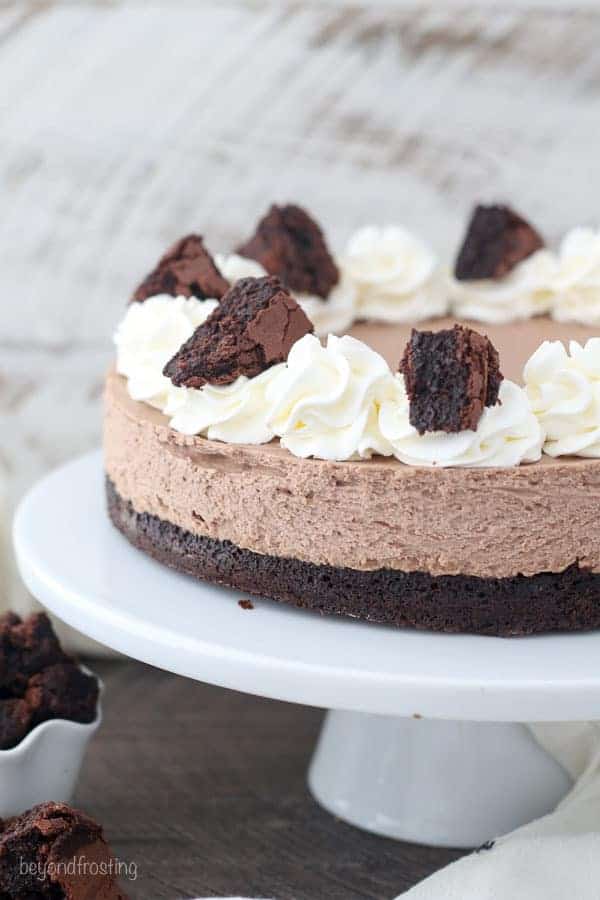 A close up of a brownie batter cheesecake on a white cake plate. The cheesecake is garnished with whipped cream and chopped brownies.