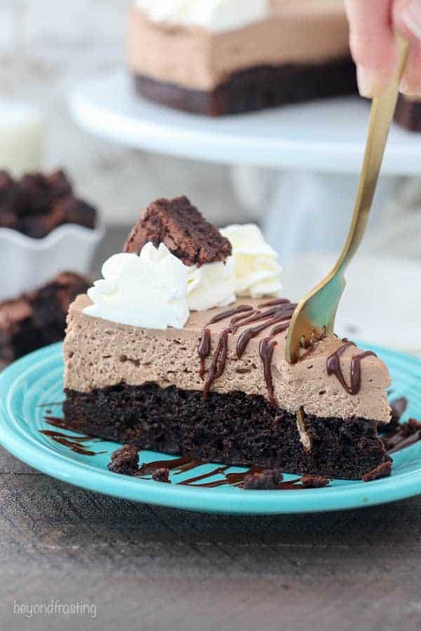 A brownie bottom chocolate cheesecake with a big gold fork digging in. It's garnished with a chocolate drizzle, whipped cream and a brownie on top.