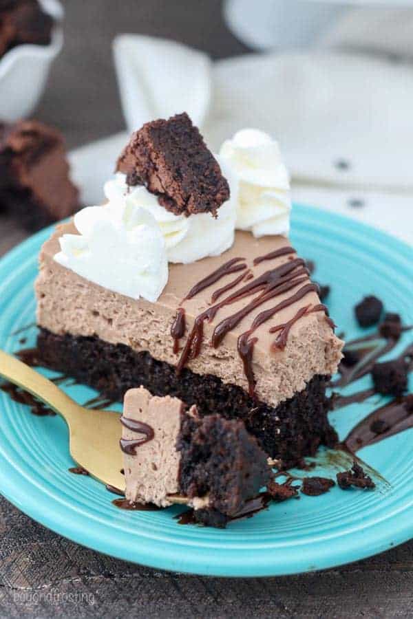 A brownie bottom chocolate cheesecake with a bite taken out of it. It's garnished with a chocolate drizzle, whipped cream and a brownie on top.