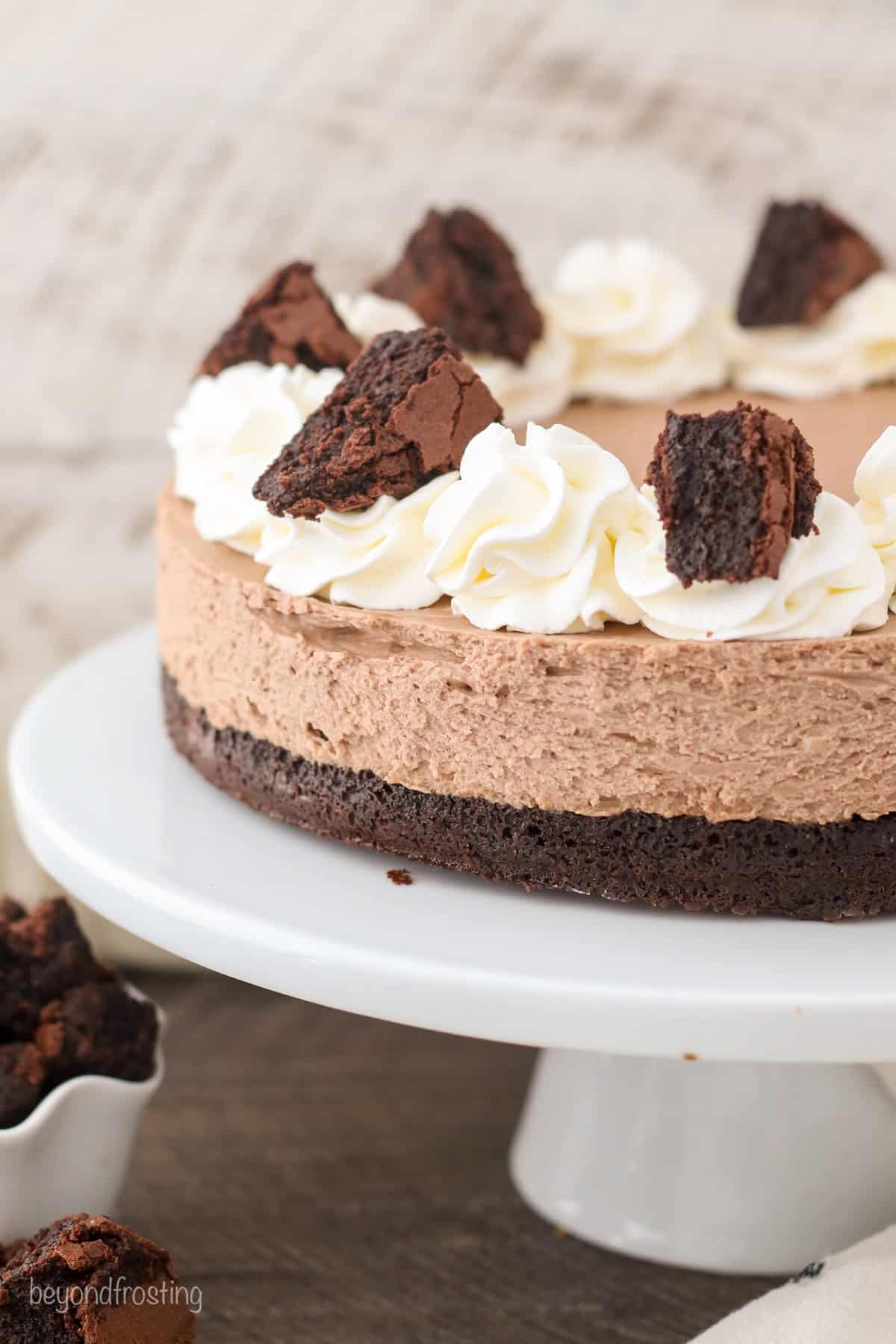 Brownie batter cheesecake topped with swirls of whipped cream and chunks of brownie on a white cake stand.