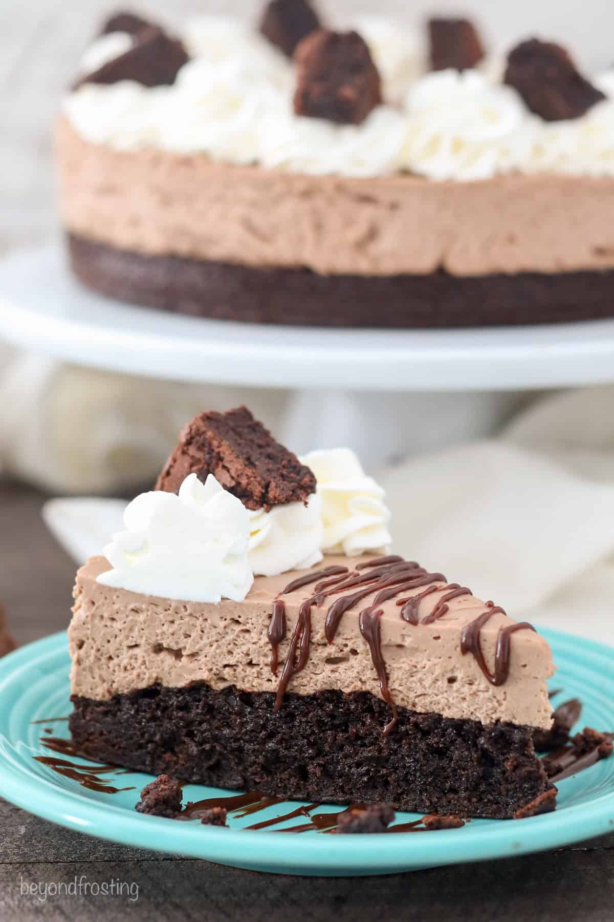 A slice of brownie batter cheesecake on a blue plate, with the rest of the cheesecake on a cake stand in the background.