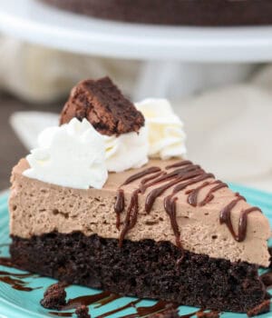 Close up of a slice of brownie batter cheesecake drizzled with hot fudge on a blue plate, with the rest of the cheesecake on a cake stand in the background.