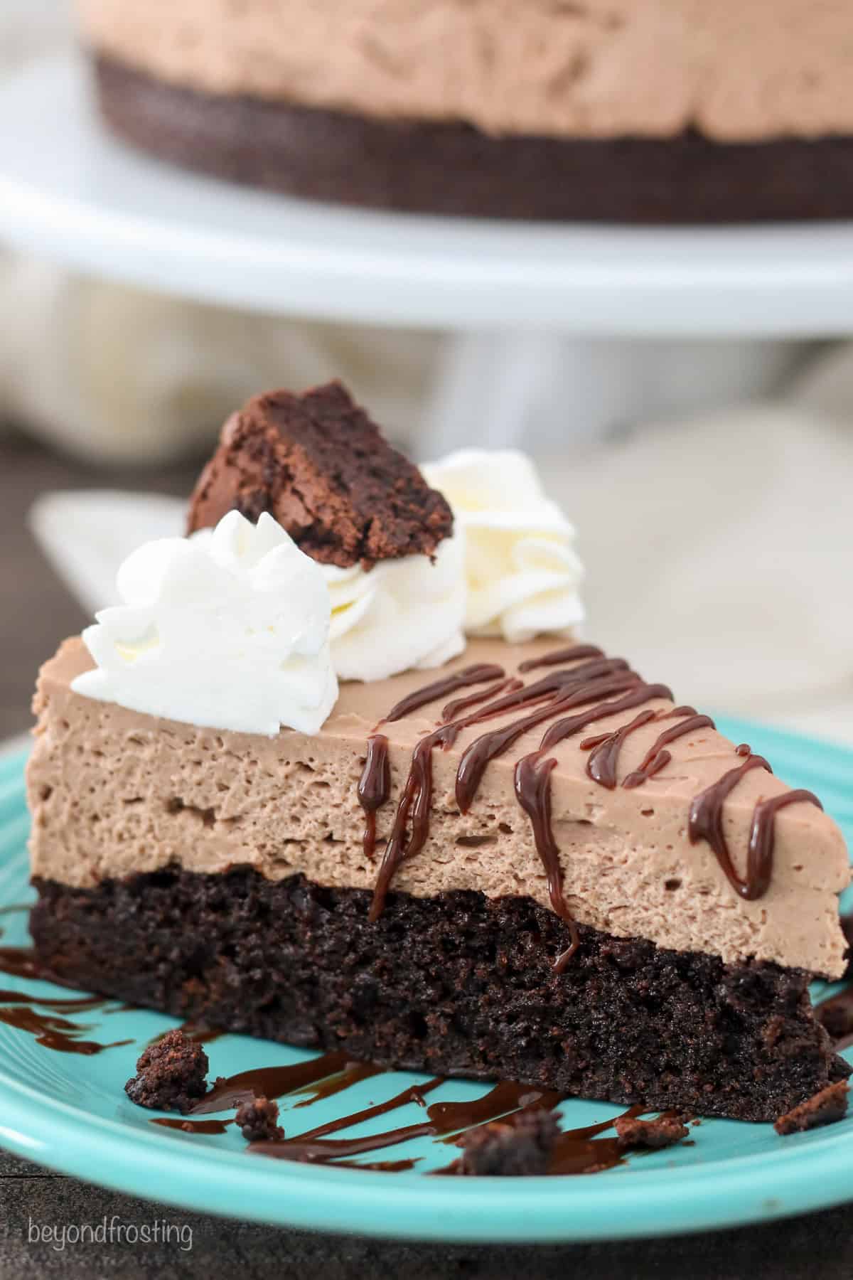 This brownie batter cheesecake has creamy no-bake cheesecake set over a thick and fudgy brownie layer. Easy to make using boxed brownie mix! #browniebatter #cheesecake Close up of a slice of brownie batter cheesecake drizzled with hot fudge on a blue plate, with the rest of the cheesecake on a cake stand in the background.