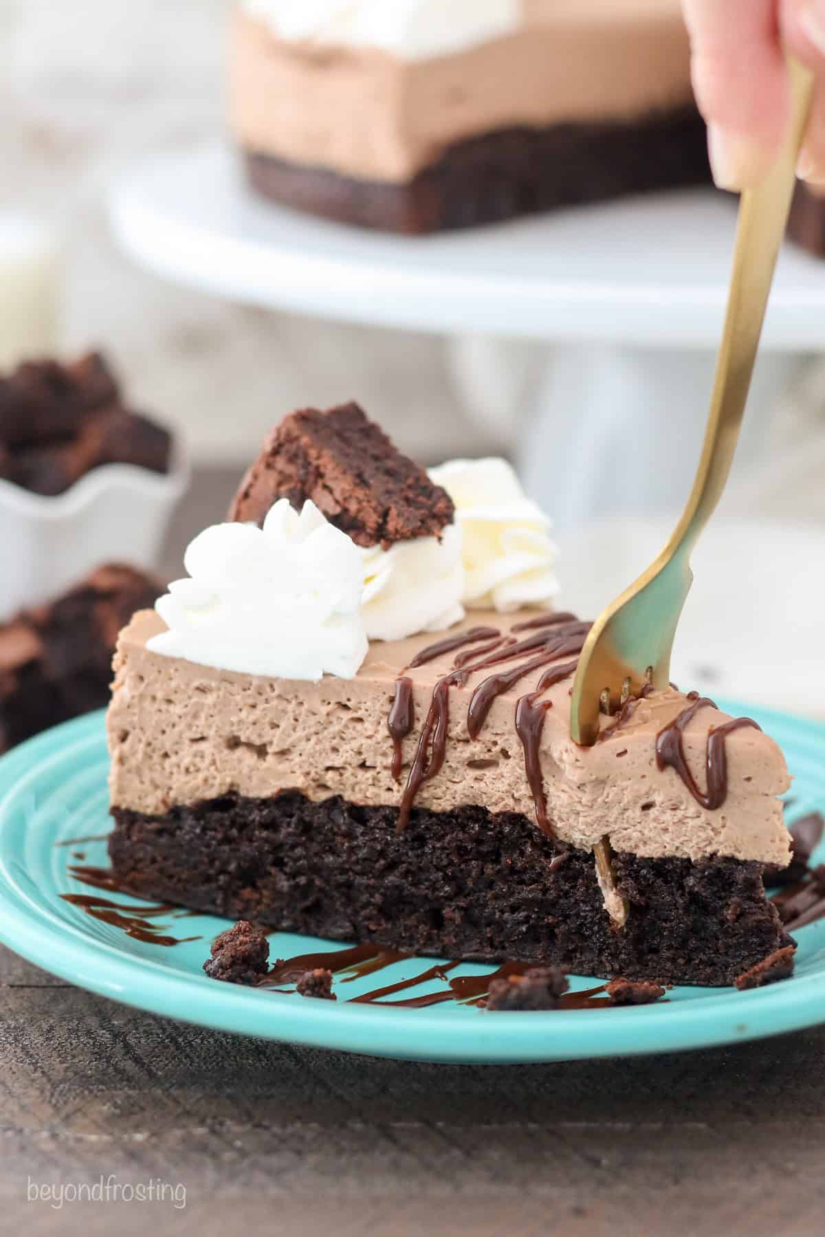 Close up of a fork stuck into a slice of brownie batter cheesecake on a blue plate, with the rest of the cheesecake on a cake stand in the background.