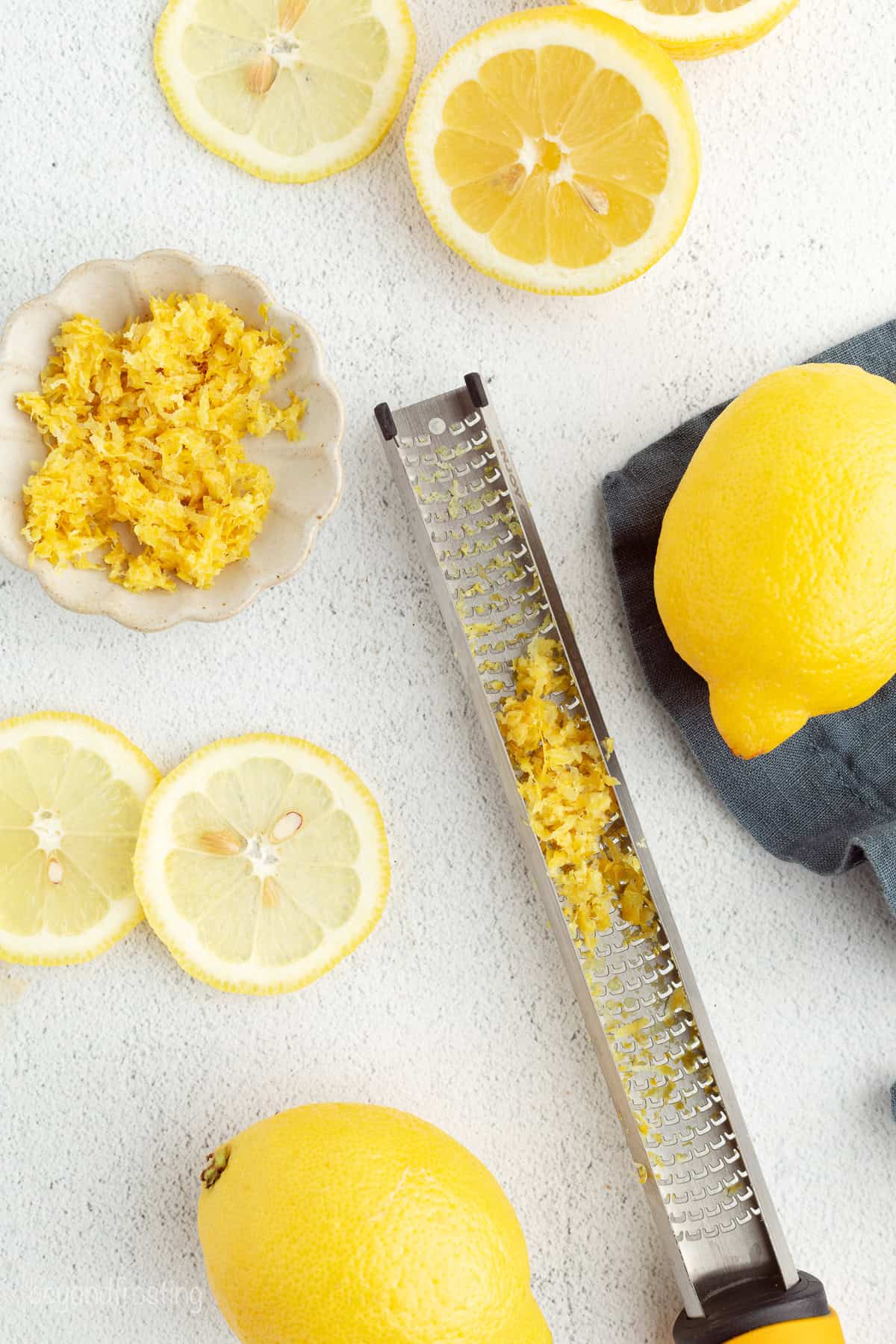 Overhead view of a lemon zester next to a bowl of zest, lemon slices, and whole lemons.