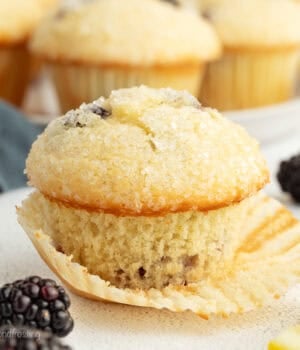 A lemon blackberry muffin partially unwrapped from a paper wrapper, with more muffins in the background.