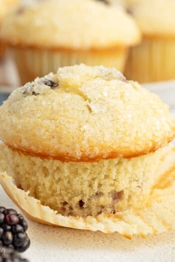 A lemon blackberry muffin partially unwrapped from a paper wrapper, with more muffins in the background.