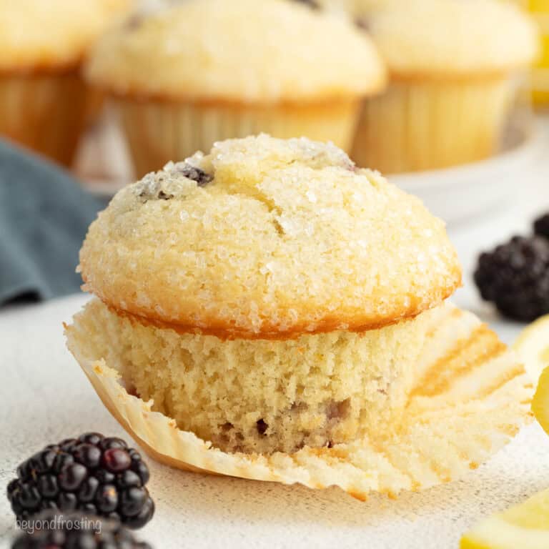 A lemon blackberry muffin partially unwrapped from a paper wrapper, with more muffins in the background.