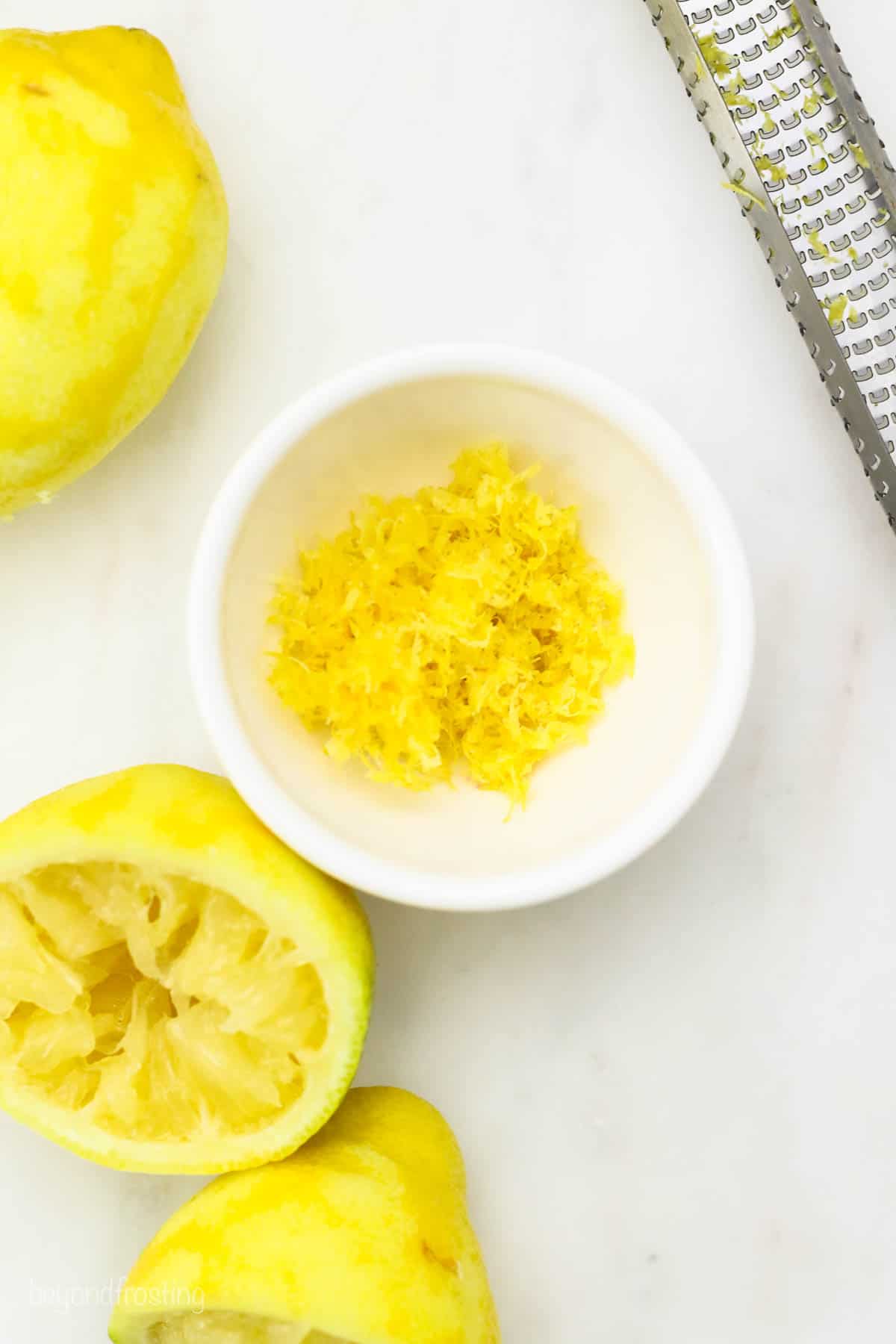A small white bowl with lemon zest next to a lemon and a microplane grater