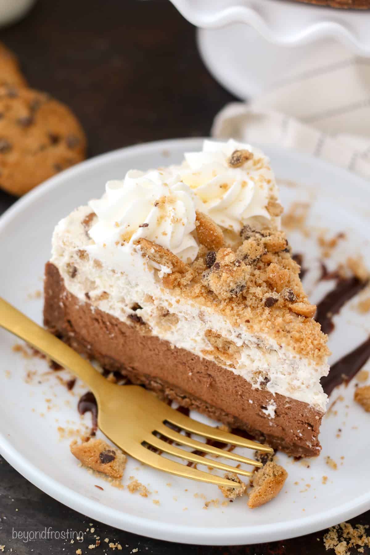 A slice of chocolate chip cookie mousse cake topped with whipped cream and crushed cookies on a plate next to a fork.