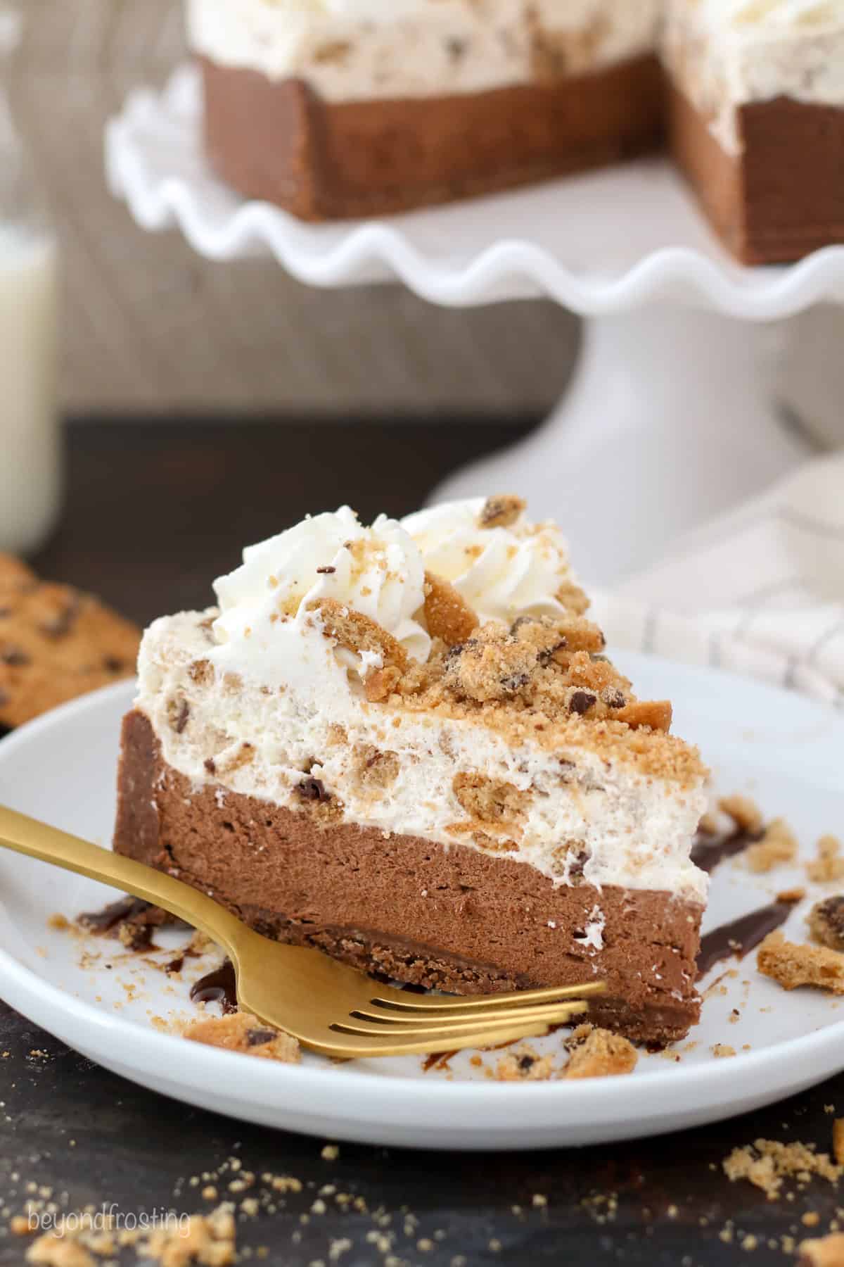 A slice of chocolate chip cookie mousse cake on a plate next to a fork, with the rest of the cake in the background.