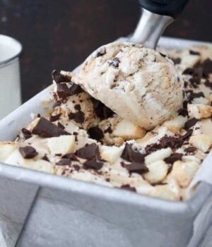 An ice cream scoop digging into a container of ice cream loaded with chocolate chunks and lady fingers.