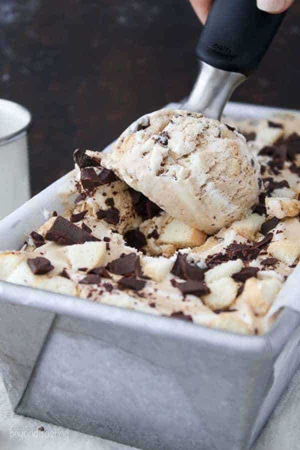 An ice cream scoop digging into a container of ice cream loaded with chocolate chunks and lady fingers.