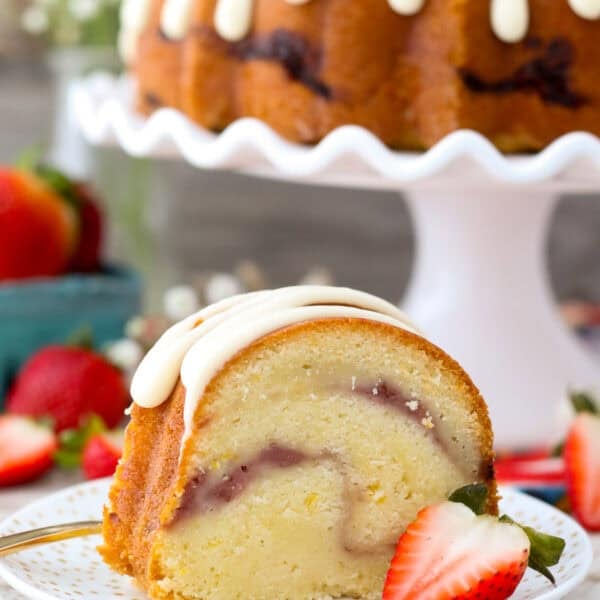 A slice of strawberry bundt cake on a white plate with the rest of the cake on a cake stand in the background.