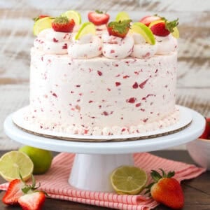 A three-tier strawberry margarita layer cake frosted with strawberry buttercream and decorated with rosettes, fresh berries, and lime wedges on a cake stand.
