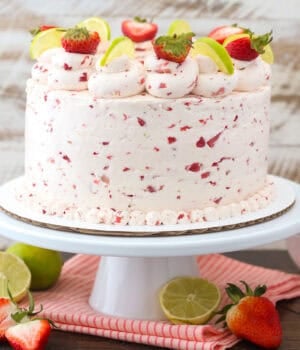 A three-tier strawberry margarita layer cake frosted with strawberry buttercream and decorated with rosettes, fresh berries, and lime wedges on a cake stand.